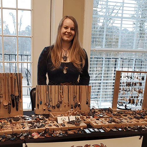Ashley Nolan smiling behind a display table with handmade crystal jewelry pieces